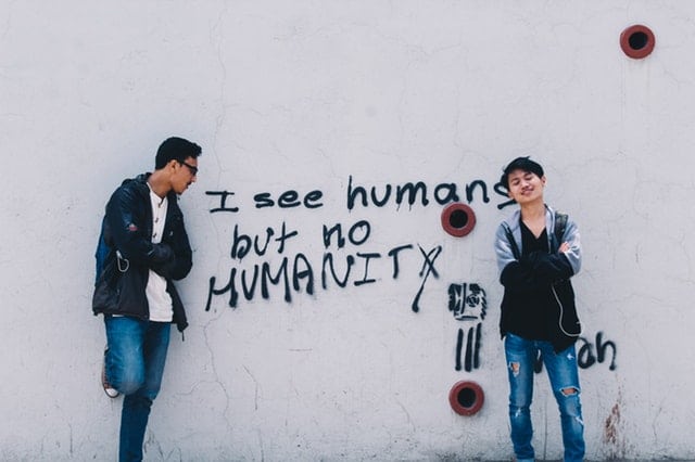 "I see humans but no humanity" writing on white wall with 2 boys standing at each side.